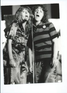 Two women sing in a theatrical manner into a microphone