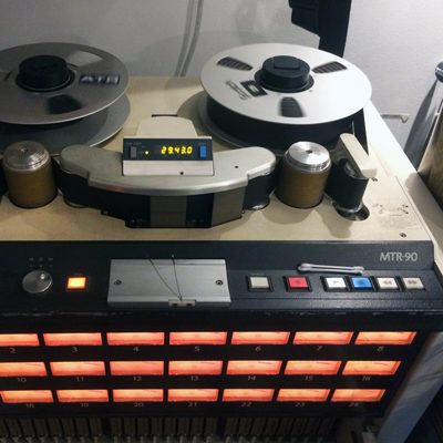chunky 2 inch tape reels spinning on flat surface of washing-machine-sized tape machine, with multiple orange-lit VU meters