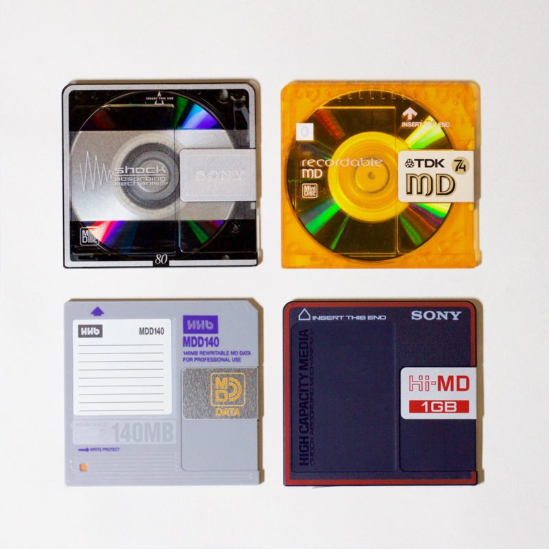 4 square plastic slim cassettes, top row: transparent and grey and transluscent amber, revealing shiny discs inside which resemble mini CDs. Bottom row both opaque, left: grey, labelled 140MB re-writable MD Data, and right: Hi-MD 1GB