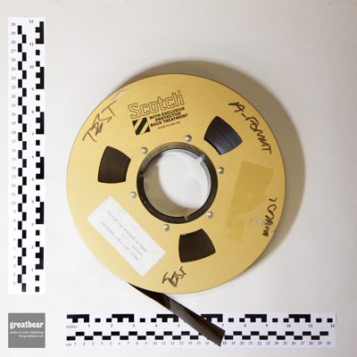 gold-coloured tape spool with dark brown / black one inch video tape, with rulers indicating 9¾ inch (24.8 cm) spool diameter