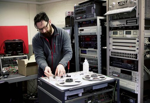 Man tending to tape machine, surrounded by racks of audio and video machines and monitoring equipment