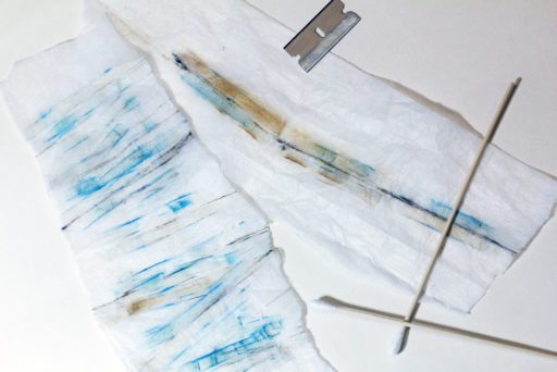 pieces of white lint cloth striped with blue, brown and black staining, a razor blade and long-stick cotton buds